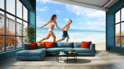 Happy beach couple vacation getaway. Young people in bikini and swimwear running holding hands together having fun on tropical beach paradise. Perfect blue ocean water and white sand. Wall mural