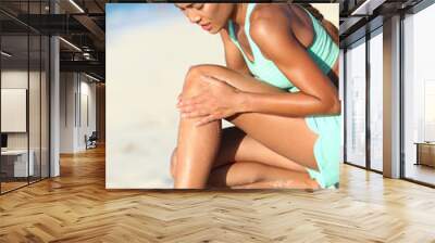 Runner injury - running woman upset with hurting knee pain. Asian female athlete with painful legs holding her leg in ache because of sport problem. Sporty girl jogging on beach. Wall mural