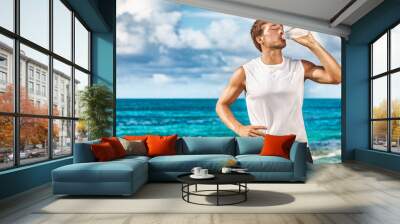 Sports drink fitness man drinking water botlle during outdoor exercise workout on beach. Dehydrated athlete runner after run sweating in summer heat exercising cardio. Wall mural