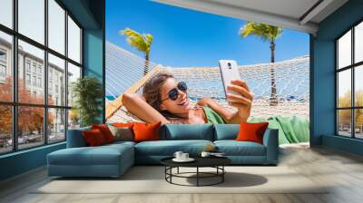 Summer beach lifestyle young woman using phone app texting on smartphone relaxing at tropical Caribbean resort on hammock. Mobile wifi internet on travel holidays. Vacation fun. Wall mural