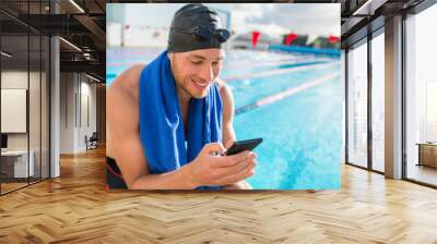 Swimmer man with swimming cap and goggles at pool using his mobile phone texting on smartphone app after training in outdoor pool. Happy athlete holding cellphone at fitness centre. Wall mural