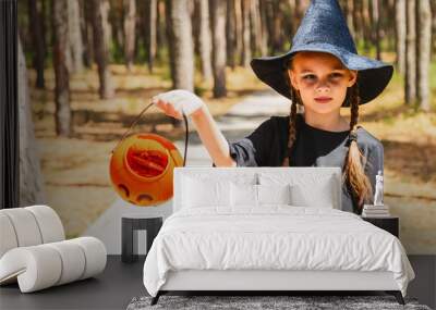 Child girl wearing witch costume holding pumpkin bucket with candies, standing in a forest on Halloween. Wall mural