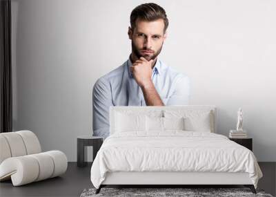 I have some doubt. Handsome young man looking at camera and keeping hand on chin while standing against white background. Wall mural