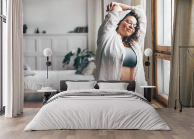 Curly haired overweight young woman in blue top and shorts with satisfaction on face accepts curvy body shape in stylish bedroom. Wall mural