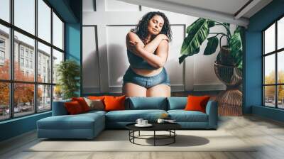 Curly haired overweight young woman in blue top and shorts with satisfaction on face accepts curvy body shape in stylish bedroom. Wall mural