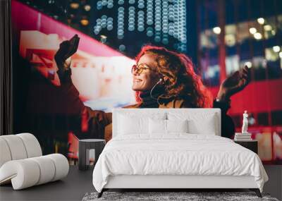 Curly haired young woman in glasses wearing light coat listens to music with headphones and makes selfie using phone camera against skyscraper in night city. Wall mural