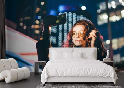 Curly haired young woman in glasses wearing light coat listens to music with headphones and makes selfie using phone camera against skyscraper in night city. Wall mural