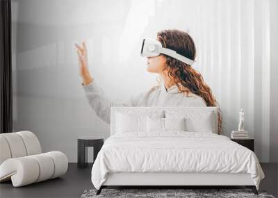 Curly haired young woman in virtual reality glasses moves hands touching online object and looks around standing in white futurisctic tunnel Wall mural