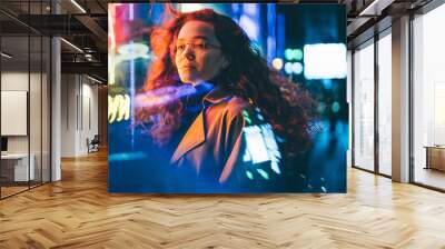 Curly haired young woman tourist with light makeup in glasses looks around standing near bar with colorful neon sign against night megalopolis Wall mural