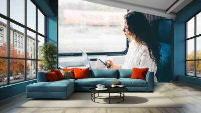 Freelancer girl working with laptop in the train. Girl looking to the phone in her hand. Business travel or technology concept. Wall mural