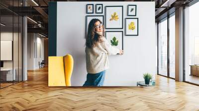 Skilled woman designer with long loose curly hair holds wooden frame with dried leaf applique and hangs on decorated wall in new apartment backside view. Wall mural