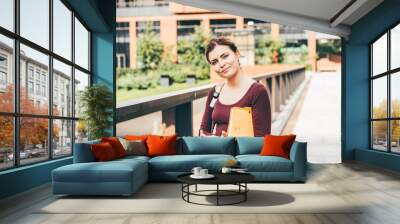 Smiling student woman in casual clothes stands holding notebooks and backpack in city street. Wall mural