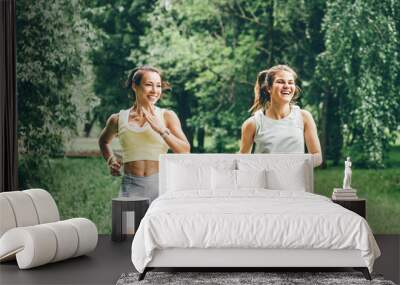 Smiling young women with ponytails in comfortable tracksuits run together along empty wooden track in green public garden on warm summer day Wall mural