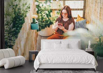 Woman wearing brown blouse and sunglasses works on modern laptop and phone siting on pillows on light terrace on sunny day closeup Wall mural