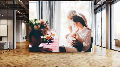 young woman breastfeeds baby and talks to grey haired mother and sitting on wooden chair. Wall mural