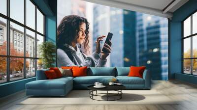 Young woman holds smartphone and flips through information on gadget screen standing against big modern financial centers in evening Wall mural