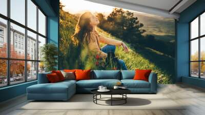 young woman traveler with long loose curly hair sits on green grass meadow with flowers and types on Wall mural