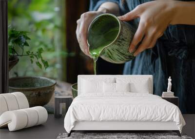 Woman in blue dress pouring matcha tea from  traditional Japanese teapot into two green bowls.  The setting is a traditional Japanese scene with plants on the table and outside.  Wall mural