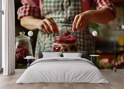 Woman preparing homemade jam with cranberries, using glass jars and fresh berries on the kitchen counter. The background is blurred to focus attention on the jar. Wall mural