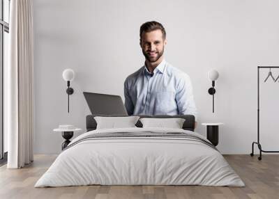 Working with joy. Handsome young man using his laptop and looking at camera with smile while standing against white background. Wall mural