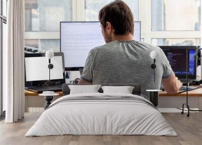 A man in a gray t-shirt is sitting at a workplace with two laptops and a monitor near the window. Remote work from home in quarantine. Back view Wall mural