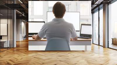 A man is sitting at a workplace with two laptops and a monitor near the window. Remote work from home in quarantine. Back view Wall mural