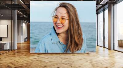 Young diverse woman in yellow glasses smiling, happy. standing by seaside Wall mural