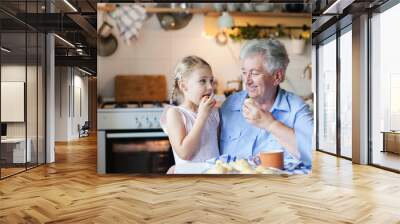 Child girl and senior woman are eating cookies in cozy kitchen at home. Happy family is enjoying handmade pastries. Grandmother and funny kid are tasting delicious food. Wall mural