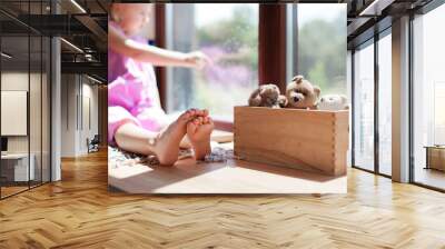 Child is sitting barefoot on window sill at home. Kid is looking out window. Little girl with wooden box of toy teddy bears. Wall mural