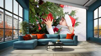 Cute child girl is decorating Christmas tree with red ornaments outside. Little kid in santa hat at winter market on town street. Cozy festive and New Year atmosphere in Dubrovnik, Croatia Wall mural