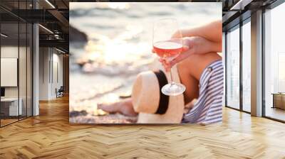 Girl is sitting on sea beach with wineglass of wine at sunset in summer vacation in resort. Tourist woman in striped dress with straw hat is enjoying life, view, traveling, relaxing, drinking. Wall mural