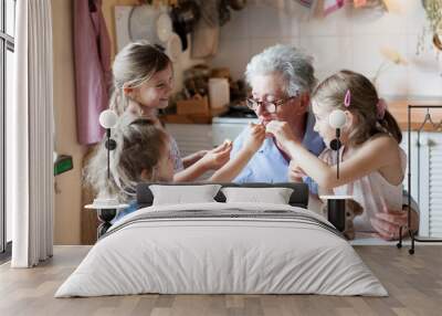 Kids treat grandmother at home. Happy family eating cookies in cozy kitchen. Senior woman and funny children tasting delicious food together, enjoying handmade pastries. Wall mural