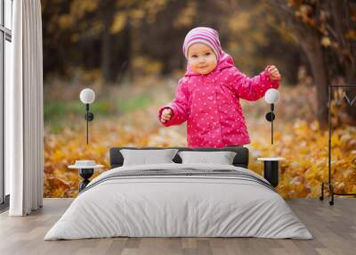 Little kid is playing and walking in autumn park. Baby looks at fallen leaves and smile. Girl is dressed in warm hat and jacket. Wall mural