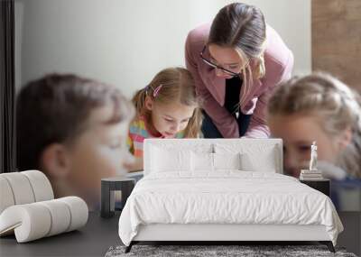 School teacher helping pupils studying, reading book. Lesson for kids in elementary college. Children sitting at desks in classroom. Woman guiding in-person, teaching primary students. Back to school Wall mural