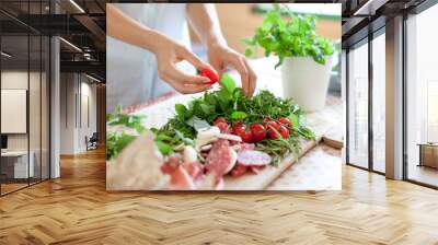 Woman is cooking in home kitchen. Female graceful hands hold cherry tomatoes and greens. Ingredients for preparing italian or french food are on table on wooden boards. Lifestyle moment. Close up. Wall mural