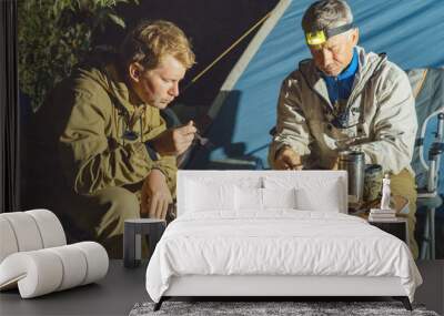 Dinner of tourists late in the evening against the backdrop of a camping tent. Two men sit on folding travel chairs at a folding travel table. There are reusable tourist dishes and thermomugs on the Wall mural