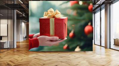 Red festive New Year gift box with a yellow bow in the hands of a child on the background of a green Christmas tree Wall mural