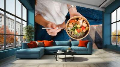 Girl holds a paper plate with healthy food sitting on the floor. Home delivery food. Healthy eating concept. Wall mural