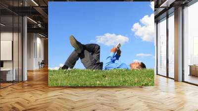 Businessman lying down on grass against blue sky with tablet. Man relaxing outdoors and working remotely Wall mural