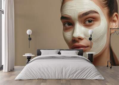 A young European lady poses with a facial mask, enjoying beauty treatments, stood over a beige studio background, and looks directly at the camera. Wall mural