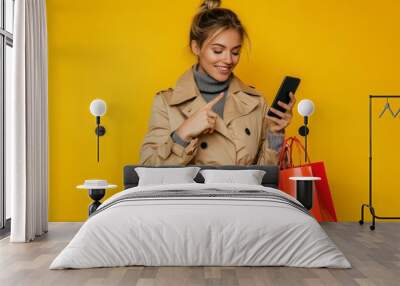 On a yellow background, a young woman in autumn clothes holds a shopping bag and a mobile phone and shows something Wall mural