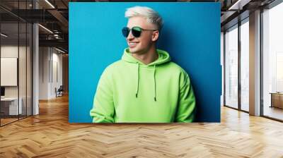 Young man smiling and having fun in trendy sun glasses and lime sweater on blue colored background - millennials have fun and smile often Wall mural