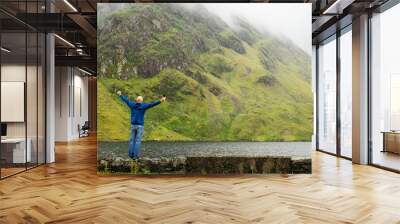 Bald excited male tourist with his hands in the air standing on a stone fence by a lake, with beautiful mountain in the backgroun. Connemara, Ireland. Concept scene of Wild Atlantic way route. Wall mural