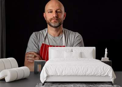 Professional butcher fishmonger wearing grey t shirt and classic red and white apron. Caucasian male in his 40s; black and grey beard, bald. Hand crossed on his chest. Black background. Wall mural