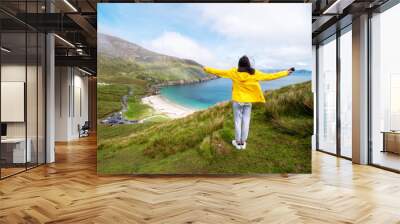 Teenager girl in yellow jacket standing on a hill and looking at stunning nature scenery of Keem bay, county Mayo, Ireland. Beautiful view on a ocean and cliffs and a beach. Warm day, cloudy sky. Wall mural