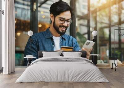 Bearded handsome man makes purchase online. A hipster wearing casual shirt holds a mobile phone and a credit card orders food. Smiling guy is using smartphone and debit bank card for paying in e-shop Wall mural