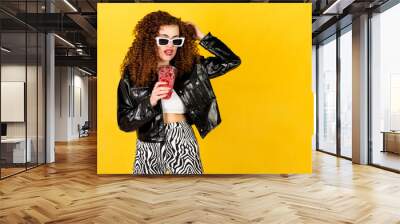 Funny and cool girl with curly hair and trendy white sunglasses in black leather jacket drinking cocktail. Isolated on yellow background. Wall mural