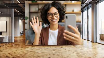 Happy woman holding smartphone and waving with hand to the camera while having video call with somebody at home . People and technologies concept Wall mural