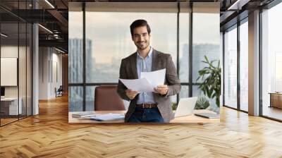 Happy young Latin business man checking financial documents in office. Smiling male professional account manager executive lawyer holding corporate tax bill papers standing at work Wall mural