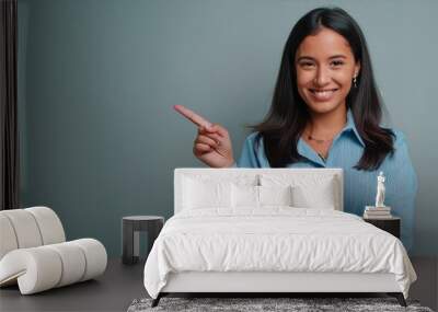 Happy young smiling professional business woman wearing blue shirt looking at camera pointing finger away at copy space showing aside presenting advertising offer standing isolated at gray background Wall mural